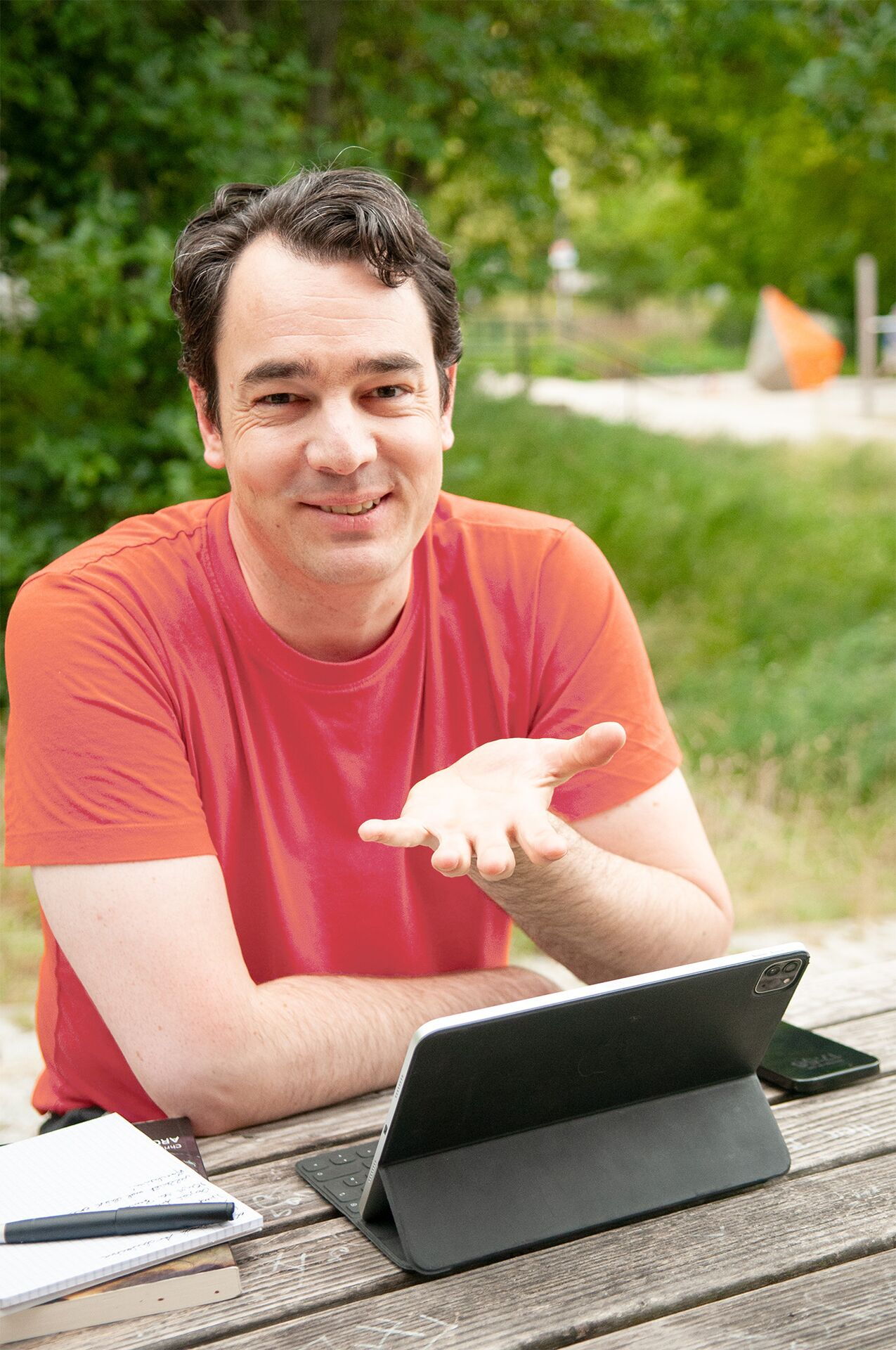 Matthias Warkus in korallrotem Shirt sitzt draußen an einem Tisch mit Tablet und Notizbuch, bereit für die Contenterstellung.