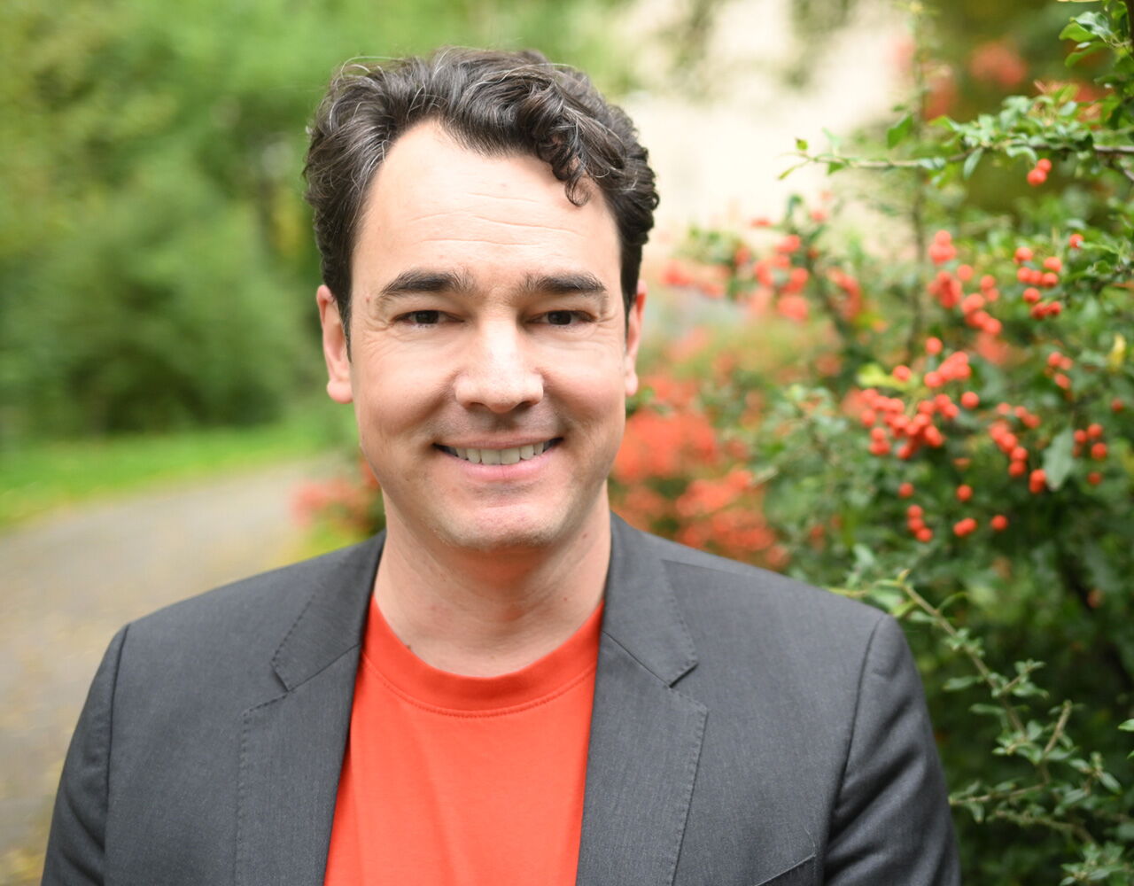 Ein Mann in rotem Shirt und dunklem Sakko steht lächelnd vor einem Busch mit roten Beeren, ideal für Contenterstellung.