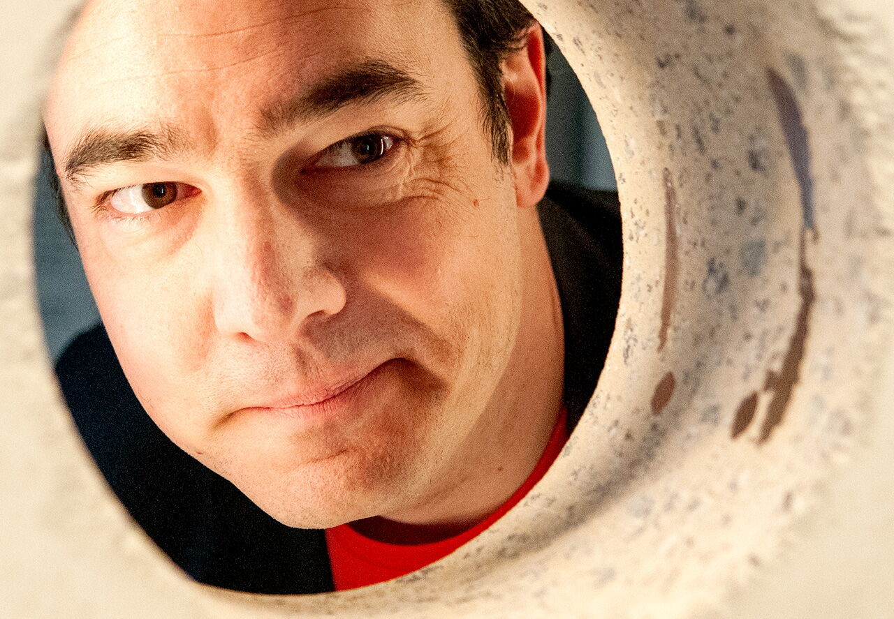 Matthias Warkus blickt neugierig durch eine runde Öffnung, was an einen kreativen Moment bei einem Science Slam erinnert.