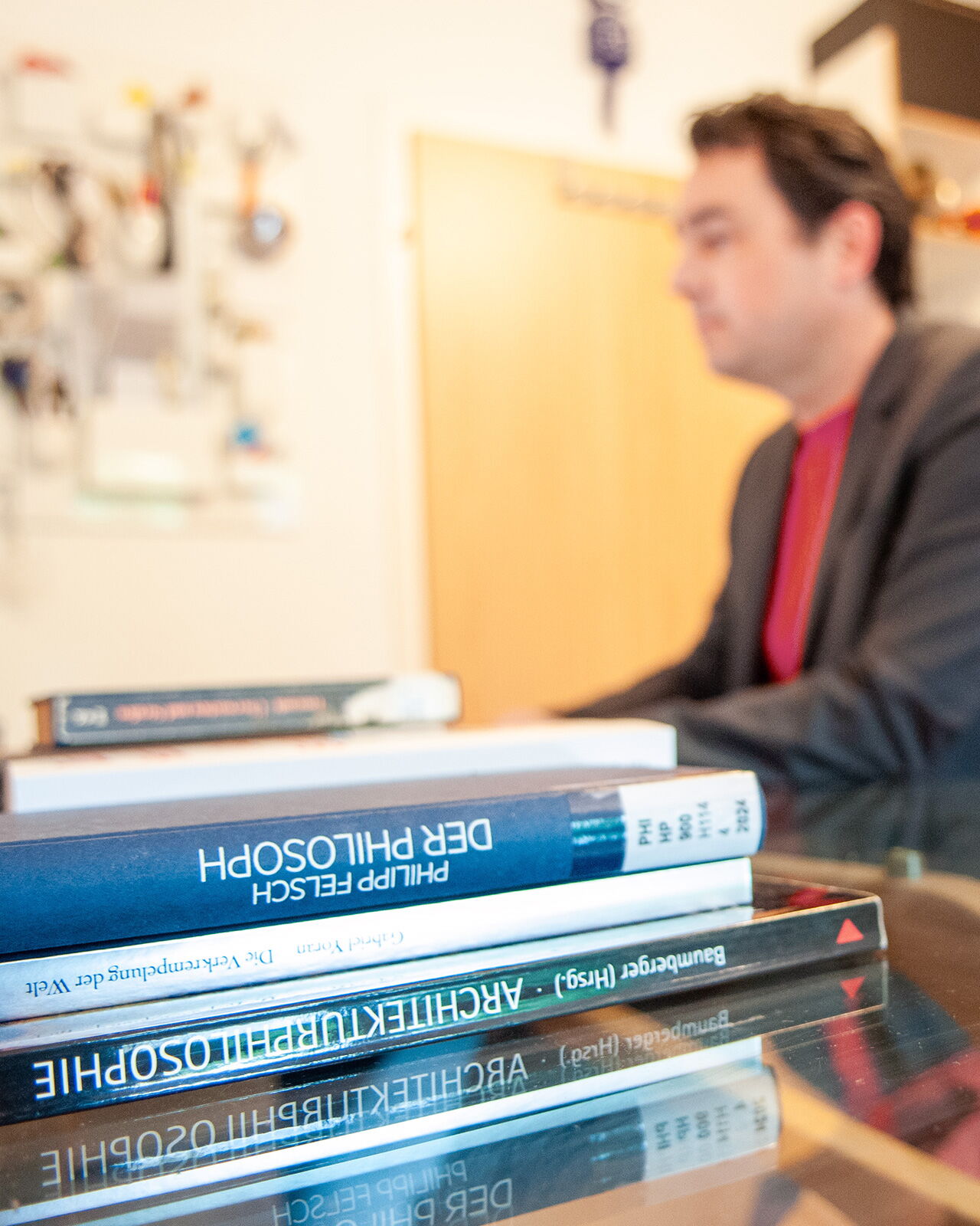 Matthias Warkus sitzt unscharf im Hintergrund, während im Vordergrund philosophische Bücher auf einem Glastisch liegen.